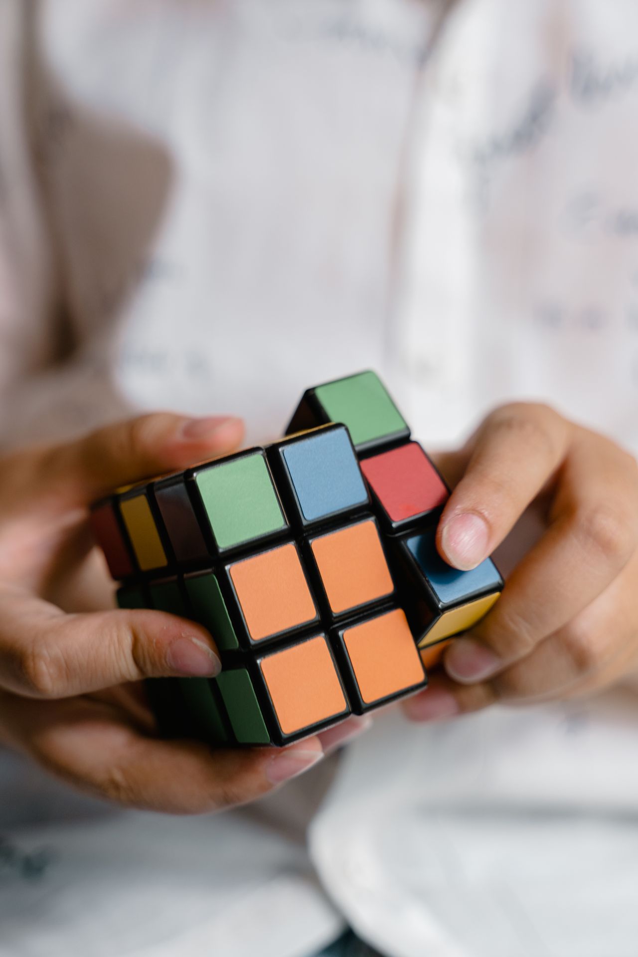 Hands holding a small puzzle cube with orange, green, blue and red tiles, similar in design to a 3x3 Rubik's Cube